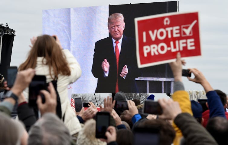 A crowd watches a man in a black suit and red tie on a large screen. Someone is holding a sign that reads 'I vote pro-life.'