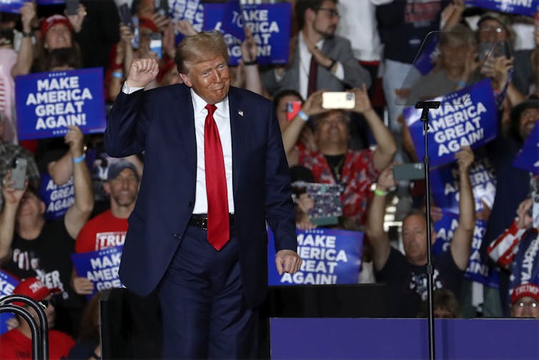 A man that is overly tanned with fluffy grey-blond hair gesturing at a campaign rally.