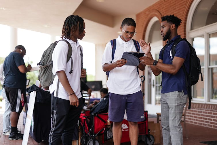 Three young men with backpacks talking; one is reading a clipboard.