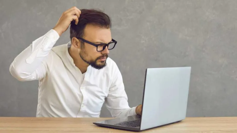 Mann mit Bart und Brille kratzt sich am Kopf und blickt verwirrt auf Laptop